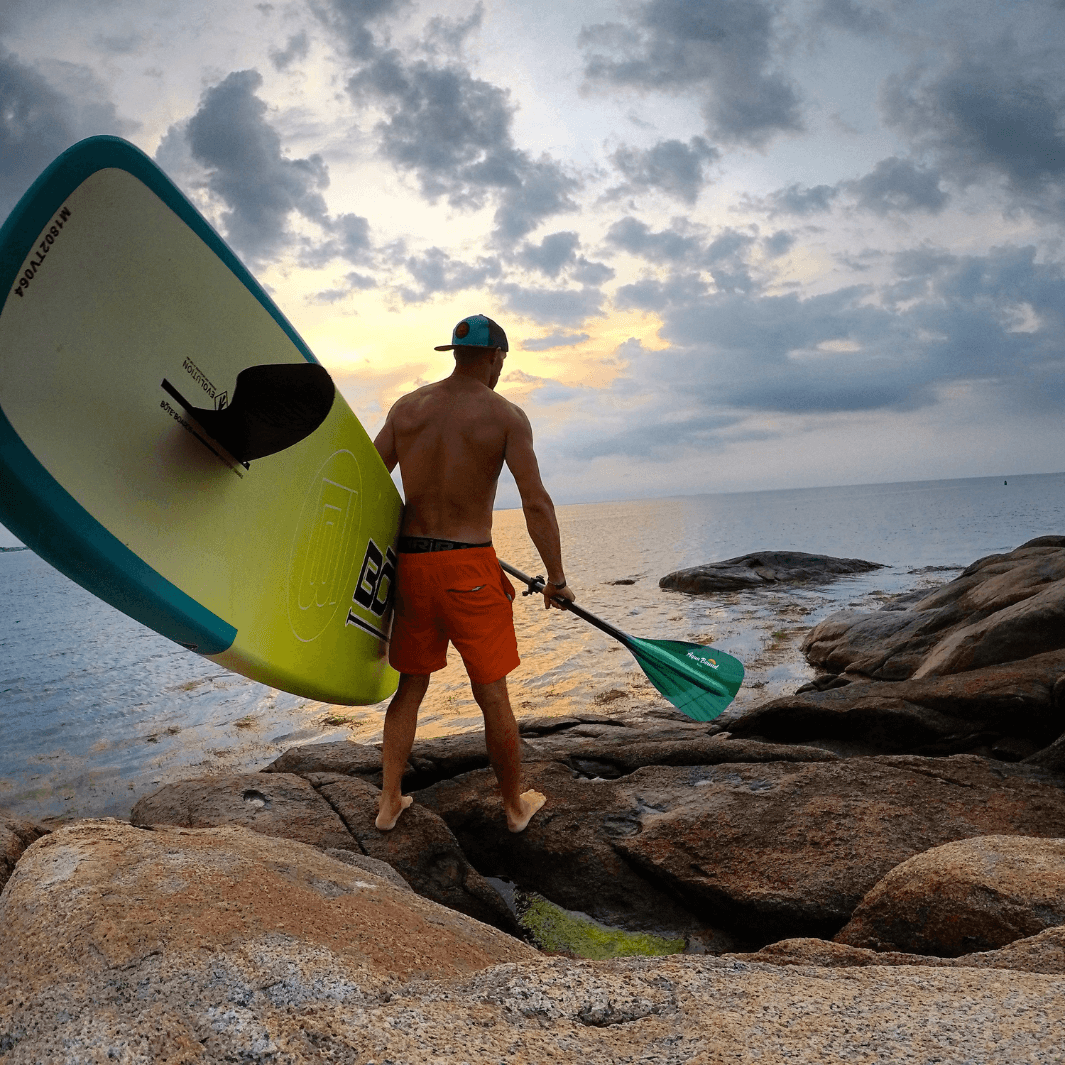 man carrying SUP board and green tide malta SUP paddle on smooth rocks next to the ocean#color-green_tide