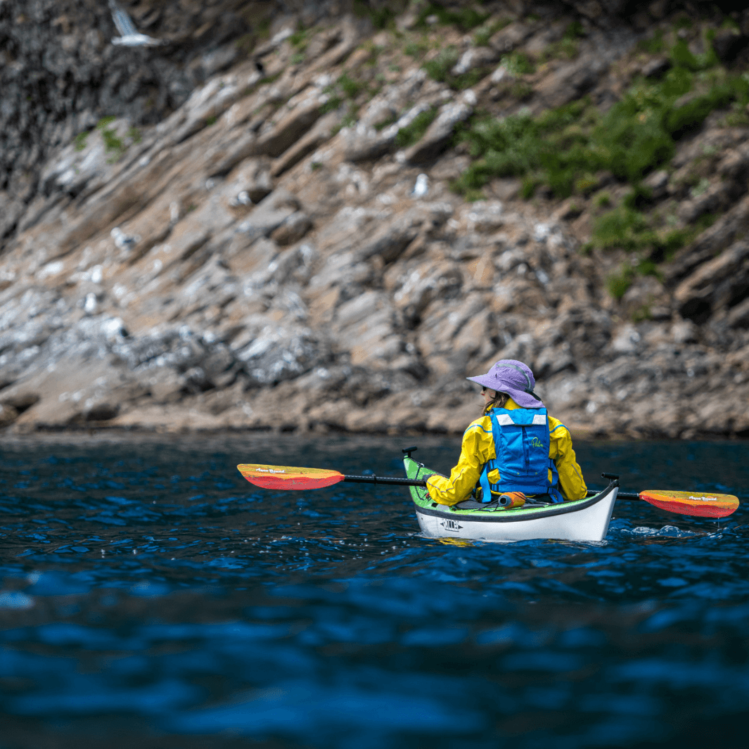 tango fiberglass 2-piece posi-lok kayak paddle fuego being paddled by female sea kayaker with rocky shoreline in background#color_fuego