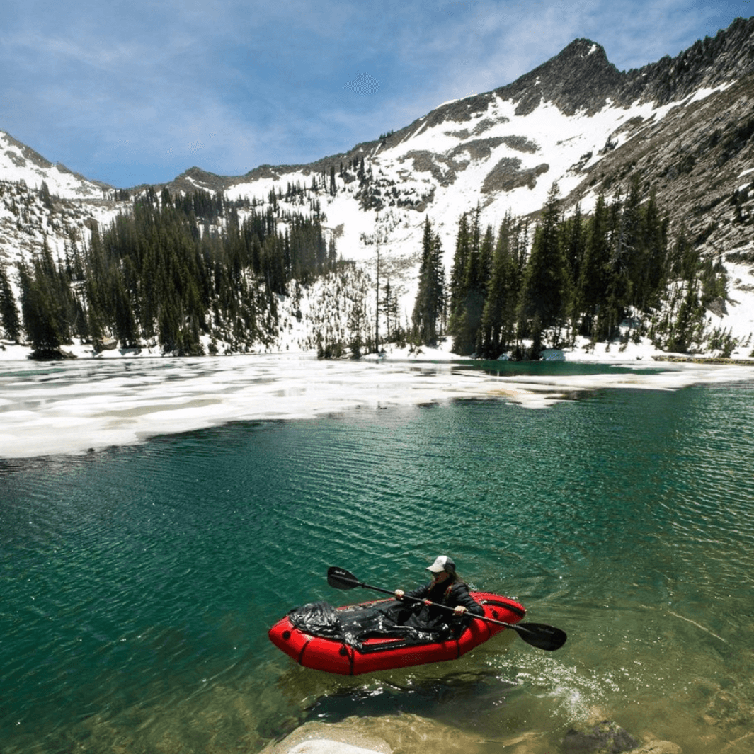 person in packraft with the whiskey carbon paddle, snowy mountains in background
