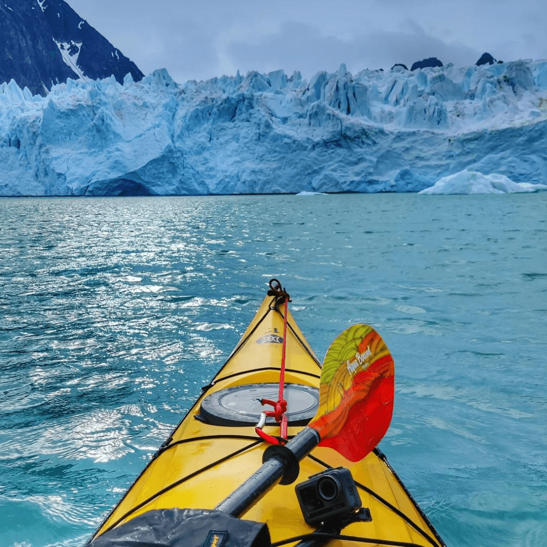 whiskey fiberglass fuego paddle across the bow of yellow sea kayak with icebergs in the background#color_fuego